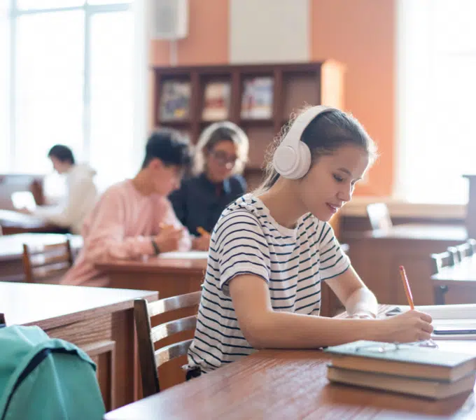 Student wearing headphones takes notes at a wooden desk while other students study in the background