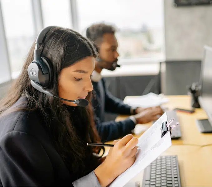 Call center employee wearing a headset writes notes while working at a computer