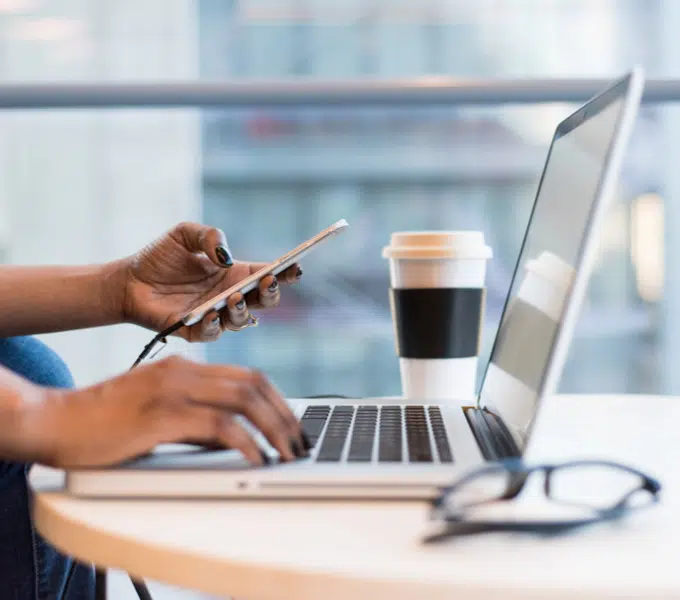 Person multitasks on a laptop and smartphone at a workspace with coffee